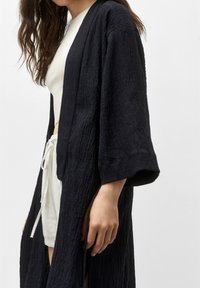 Capa exterior negra de estilo kimono con textura, de mangas anchas, que presenta un corte relajado y un sutil patrón arrugado. Llevado sobre una blusa ligera y pantalones cortos.