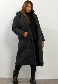 Długi czarny płaszcz puchowy z kapturem, wykonany z materiału pikowanego. Posiada zapięcie na zatrzaski i zamek błyskawiczny, kieszenie boczne oraz luźny krój.