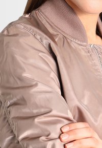 Ljusrosa satinbombarjacka med en ribbad krage, detaljerad stygn och en mjuk textur, som visar ärmen och en handdetalj.