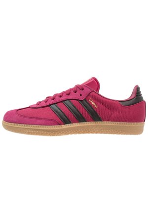 Baskets rouges Adidas Samba à profil bas avec des bandes noires, semelle en gomme et texte doré "SAMBA" sur le côté, montrées de côté extérieur.