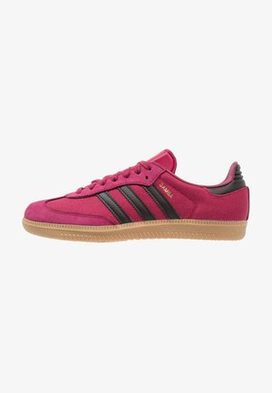 Niskoprofilowy czerwony sneaker Adidas Samba z czarnymi paskami, gumową podeszwą i złotym tekstem "SAMBA" po boku, pokazany od zewnętrznej strony.