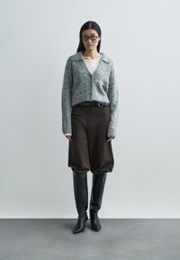 Grijze mohair cardigan met een V-hals, gecombineerd met bruine volumineuze broek en zwarte kniehoge laarzen. Eenvoudig ontwerp, minimale accessoires.