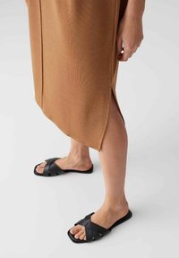 Braunes geripptes Kleid mit seitlichem Schlitz, kombiniert mit schwarzen Sandalen mit Kreuzriemen und flacher Sohle. Fokus auf Textur- und Farbkontraste.