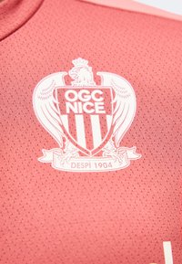 Έμβλημα του ποδοσφαιρικού συλλόγου OGC Nice με αετό και ασπίδα πάνω σε υφασμάτινο ροζ αθλητικό ύφασμα με υφή.