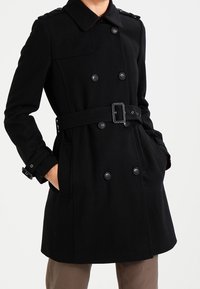 Zwarte dubbelgeknoope trenchcoat met een tailleband, grote knopen en schouderepauletten. Glad stof met een klassiek ontwerp.