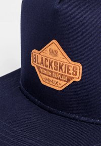 Sötétkék sapka texturált anyagból, amelyen egy kiemelkedő barna bőr címke található, rajta a "BLACKSKIES" felirattal és dekoratív kiegészítőkkel.