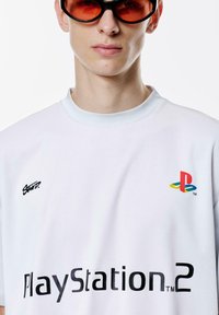 Bijela majica, teksturirani materijal, s crnim "Swoop" logom, "PlayStation 2" u podebljanim slovima i višebojnim PlayStation logom na prsima.