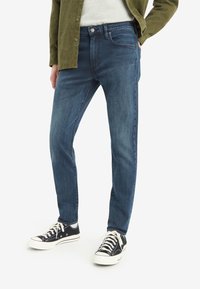 Denim jeans in dark blue with a slim fit, featuring five pockets and subtle whiskering. Paired with black canvas sneakers.