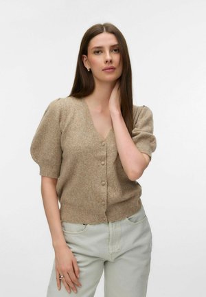 Mladá žena s dlouhými hnědými vlasy, oblečená v béžovém krátkém svetru na knoflíky a světle šedých kalhotách, dotýkající se krku jednou rukou.