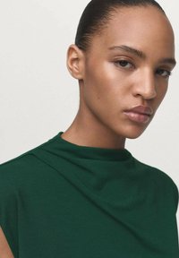 Prenda verde de cuello alto con textura suave, que presenta un diseño drapeado en el escote y hardware mínimo. Primer plano de la cara, fondo neutral.