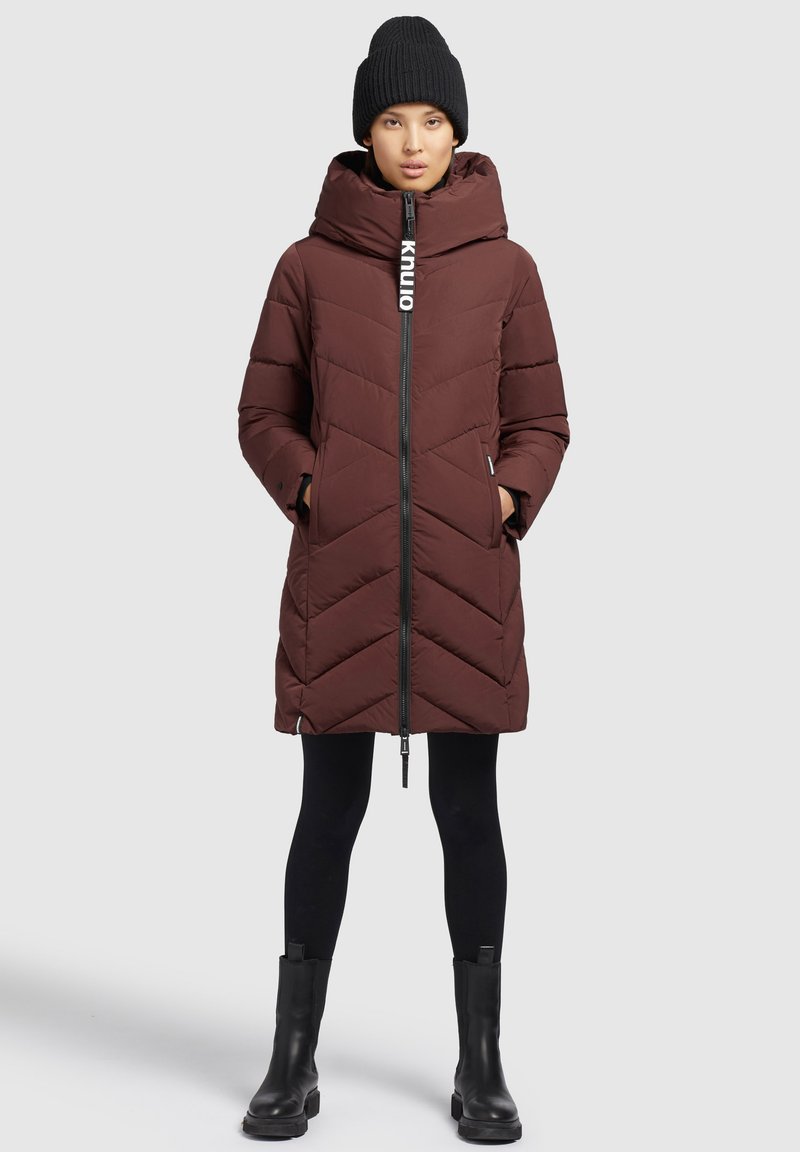 Brun vadderad jacka med chevronmönster, hög krage och dragkedja. Matchas med svarta leggings och klumpiga svarta stövlar, toppad med en stickad mössa.