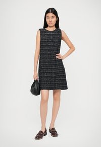 Noor naine seisab käsi puusal, kandes ilma varrukateta ruudulist tumedat kleiti, hoides musta käekotti ja pruune mokassине valge taustaga.