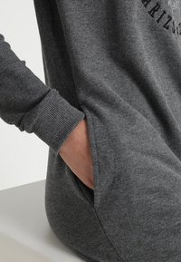 Main dans la poche latérale d'une robe-pull en maille grise avec poignets côtelés et texte partiel imprimé sur la poitrine.