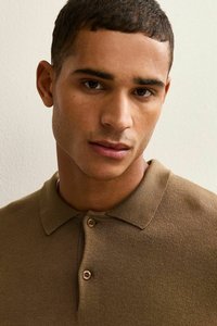 Brown polo shirt made of textured knit fabric, featuring a collar and two-button placket. Close-up of the shirt worn by a person.