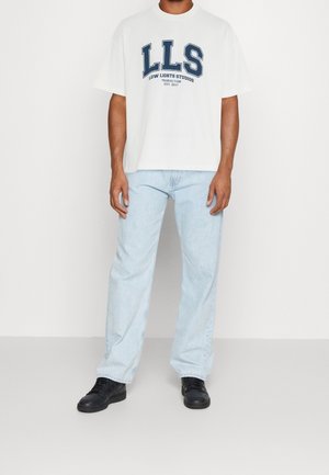 Hvid oversize t-shirt med marineblåt grafisk tryk "LLS LOW LIGHTS STUDIOS," kombineret med lyseblå denimjeans og sorte sneakers. Enkel design.