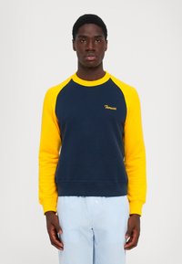 Mann iført navy blå og gul langermet hettegenser med "Fiorucci"-logo, kombinert med lyseblå og hvite stripete bukser mot hvit bakgrunn.