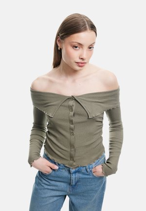 Olijfgroene geribbelde langemouwen top met off-the-shoulder ontwerp, knopen aan de voorkant en gegolfde zoomdetails. Gecombineerd met lichtblauwe jeans.