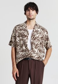 Camicia a maniche corte beige con motivo di foglie di palma marroni. Presenta una vestibilità rilassata e colletto aperto, abbinata a pantaloni marroni scuri.