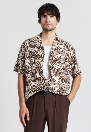Camicia a maniche corte beige con motivo di foglie di palma marroni. Presenta una vestibilità rilassata e colletto aperto, abbinata a pantaloni marroni scuri.