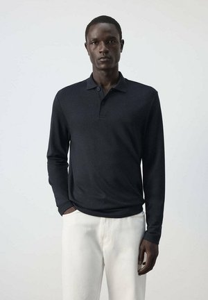 Joven de pie con una camisa polo negra de manga larga y pantalones blancos frente a un fondo liso gris claro.