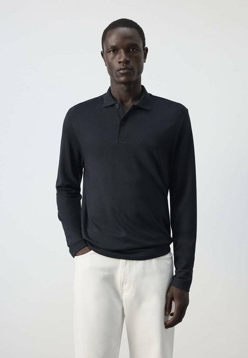 Jeune homme debout portant un polo noir à manches longues et un pantalon blanc sur un fond uni gris clair.