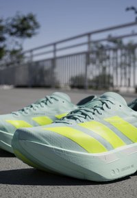 Jasnozielone buty do biegania z jaskrawymi żółtymi paskami, umieszczone na szarej nawierzchni obok metalowej poręczy i rozmytych drzew w tle.