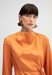 Oranje satijnen jurk met een hoge halslijn en lange mouwen, getailleerde taille met een strik. Model draagt gouden bladeren oorbellen, steil donker haar.