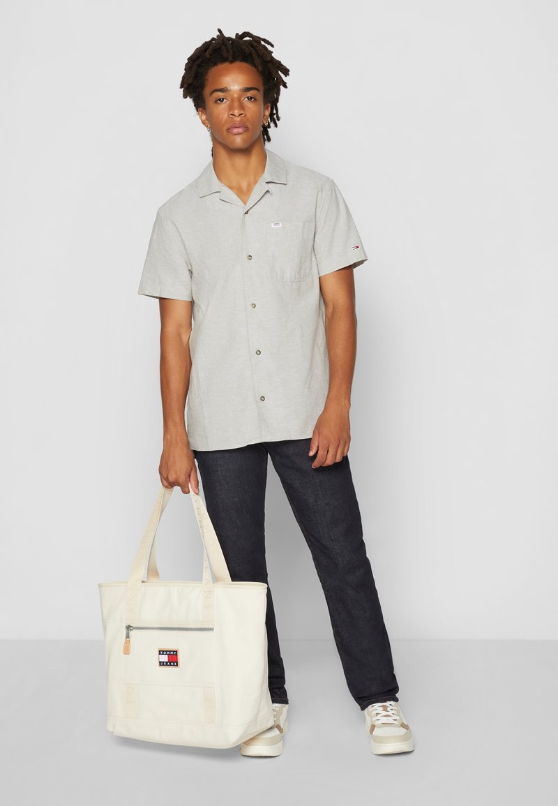 Jonge man in donkere jeans en een lichtgrijze korte mouwen shirt, met een beige tote bag in de hand, staande tegen een effen witte achtergrond.