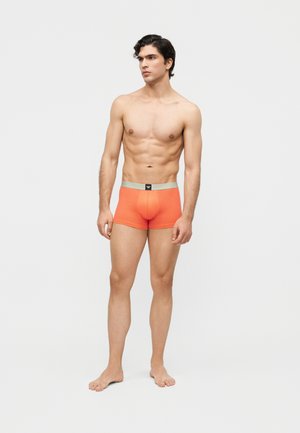 Sobiv noormees seisab paljajalu, kandes oranže bokserpükse halli vöökohaga, vaadates kergelt paremale, taust on ühevärviline valge.