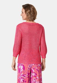 Gestrickter Pullover in leuchtendem Pink mit lockerem, offenem Muster und drei Viertelärmeln. Über Blumenmuster-Hosen getragen.