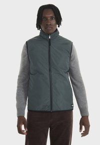 Gilet matelassé de couleur vert foncé avec un col haut et une fermeture éclair à l'avant, porté sur une chemise à manches longues gris clair, associé à un pantalon en velours côtelé marron.