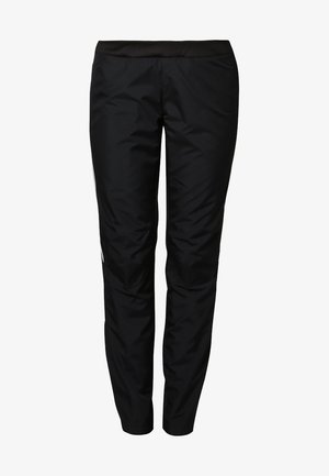 Femme portant un haut de sport noir sans manches, un pantalon noir et des baskets noires, debout devant un fond clair uni.