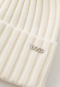 Căciulă tricotată de culoare crem, cu o textură ribbed și un accent de logo metalic argintiu. Oferă o potrivire strânsă și un aspect moale.