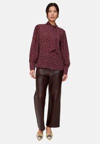 Bordeauxrote Bluse mit weißen Polka-Dots, Schleifendetail am Kragen; kombiniert mit dunkelbraunen, weiten Lederhosen und beigen spitzen Schuhen.