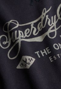 Tissu bleu marine avec texte cursif blanc « Superdry » et graphique de ciseaux avec « 1954 » sur une étiquette, faisant partie d'un logo au style vintage.