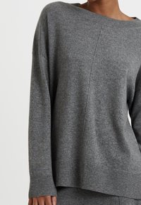 Pull en tricot gris avec un large décolleté, des épaules tombantes et des poignets et ourlet côtelés. Texture douce et silhouette décontractée.
