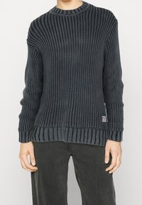 Pull en tricot bleu foncé avec texture côtelée, col rond et ourlet rayé contrastant. Doté d'un petit patch logo sur le devant.