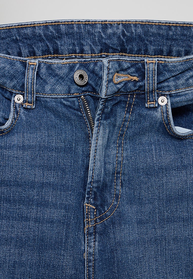 Jean en denim de teinte bleue, avec une fermeture à glissière et un bouton, cinq poches, des coutures jaunes, et des passants de ceinture à la taille.