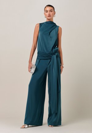 Kvinde iført teal ærmeløs buksedragt med vide ben og lang taljebånd, holder sølv clutch, accessoriseret med sølv øreringe og armbånd.