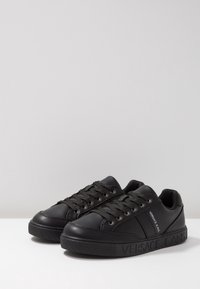 Baskets basses noires Versace Jeans avec lacets et logo embossé sur le côté et la semelle, posées sur une surface blanche.