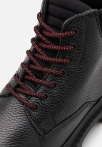 Schwarze Lederstiefel mit strukturiertem Obermaterial, roten und schwarzen Schnürsenkeln, gepolstertem Schaft und schwarzen Ösen. Verfügt über eine Gummisohle und gestickte Details.