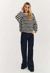 Pruhovaný svetr s plyšovou texturou v krémové a námořnické modré, doplněný tmavě modrými džíny s rozšiřujícími se nohavicemi a černými nazouvacími botami.