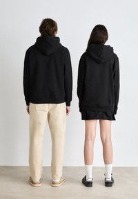 Sudaderas con capucha negras y puños acanalados, combinadas con pantalones beige y una falda negra. El calzado del modelo incluye zapatos de suela gruesa y calcetines blancos.