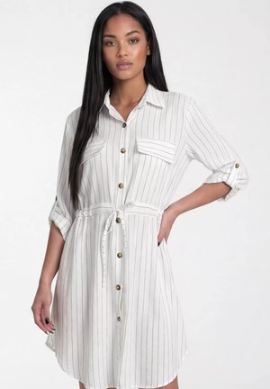 Kobieta w białej sukience zapinanej na guziki z cienkimi czarnymi pionowymi paskami, z podwiniętymi rękawami, kieszeniami na piersiach i paskiem w talii, stoi z jedną ręką na biodrze.