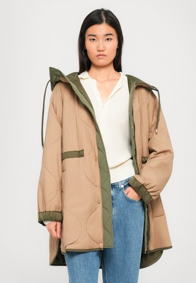Young woman wearing oversized beige and olive quilted coat over white knit top and blue jeans, standing against a plain background.
