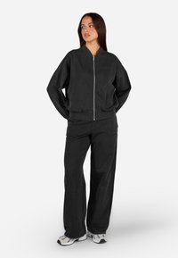 Čierna bavlnená zipsová bunda a zodpovedajúce nohavice majú voľný strih, rebrované manžety a viditeľné švy, v kombinácii so svetlými teniskami.