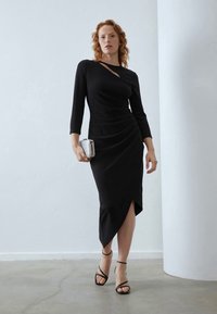 Schwarzes Kleid mit langen Ärmeln, asymmetrischem Schnitt und Raffungselementen. Kombiniert mit schwarzen Riemchenschuhen und einer kleinen silbernen Clutch.