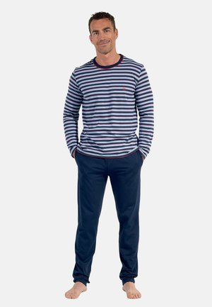 Ein Mann steht barfuß vor einem einfarbigen Hintergrund, trägt ein langärmliges blau-weiß gestreiftes Hemd und dunkelblaue Hosen.