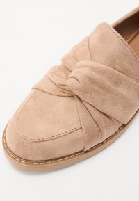 Pantofii slip-on din suede bej deschis prezintă un detaliu legat în față, vârful rotund și o talpă din cauciuc cusută.