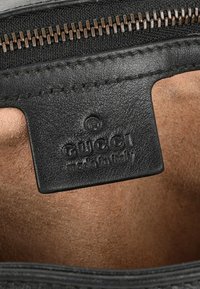 Gros plan d'une étiquette Gucci en cuir noir cousue à l'intérieur d'un sac doublé en daim sous une fermeture éclair en bronze.